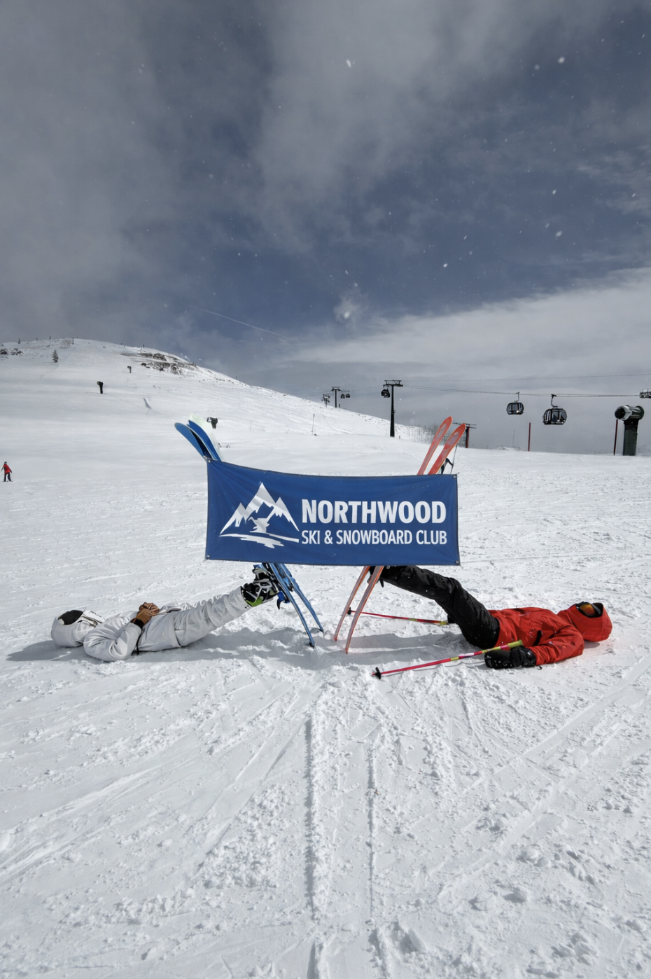 Two skiers lie on their backs in the snow with skis crossed over a blue "Northwood Ski and Snowboard Club" sign, while a ski lift and snowy slope create the perfect winter backdrop.